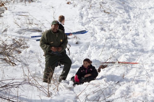 A park ranger stands in the snow with a child sitting nearby, while another child plays in the background with skis.