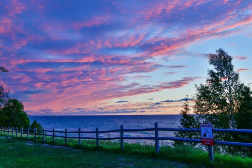 A vibrant sunset over a lake, with colorful clouds and a warning sign near a wooden fence.