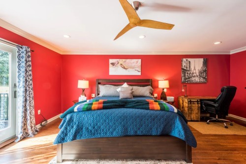 Cozy bedroom with red walls, a large bed, desk, and a ceiling fan, featuring colorful bedding and natural light.