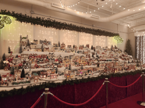 A festive display of miniature houses on a snowy hillside, adorned with lights and surrounded by holiday decorations.