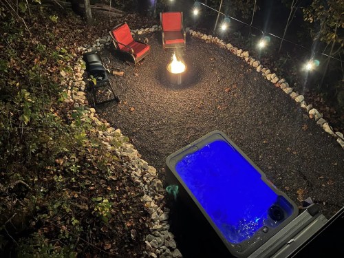 A cozy outdoor space with a hot tub, fire pit, and chairs, surrounded by stones and string lights in a wooded area.