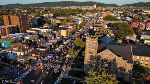 Aerial view of a bustling street festival with colorful tents and crowds, surrounded by buildings and greenery.