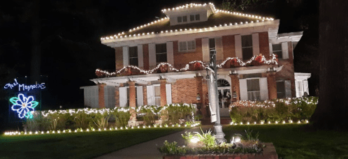 A beautifully lit brick house decorated with string lights and festive displays, surrounded by greenery at night.