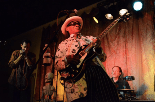 A musician in a white hat and patterned shirt plays guitar on stage, accompanied by a harmonica player and drummer.