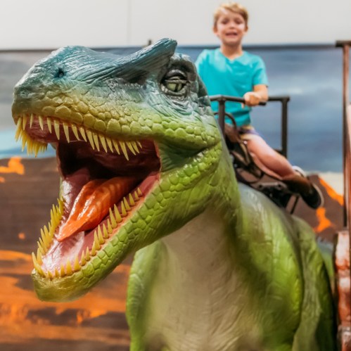 A child smiles while riding a large dinosaur statue with an open mouth and vibrant colors.