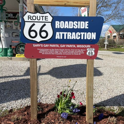 Sign for Route 66 Roadside Attraction in Gay Parita, Missouri, with flowers in front and vintage gas station in background.