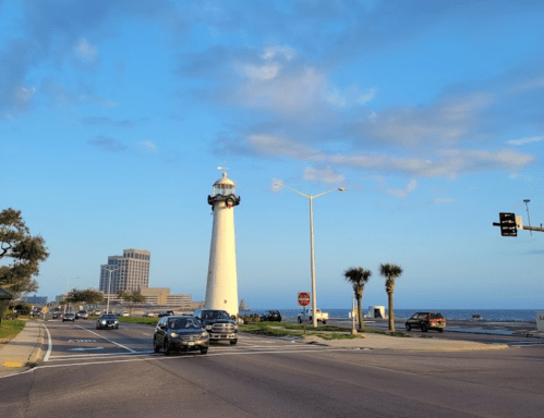 A lighthouse stands by the road near the ocean, with cars passing and palm trees lining the street under a blue sky.