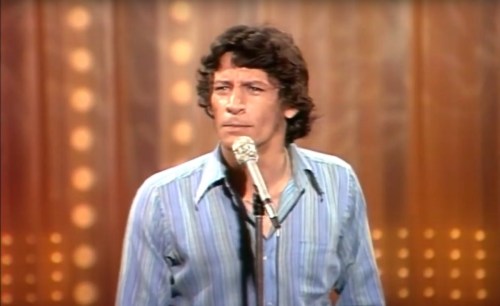 A man with curly hair stands at a microphone, performing on stage with a wooden backdrop and bright lights.