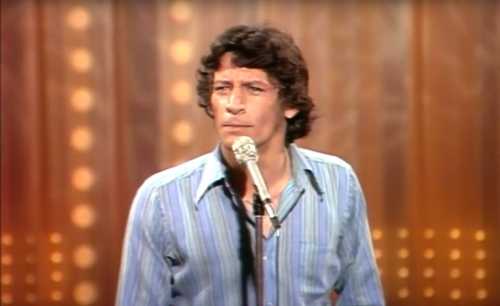 A man with curly hair stands at a microphone, performing on stage with a wooden backdrop and bright lights.