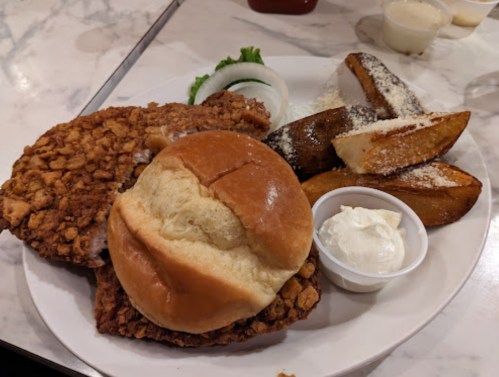 A plate with a fried chicken sandwich, seasoned potato wedges, and a small cup of dipping sauce.