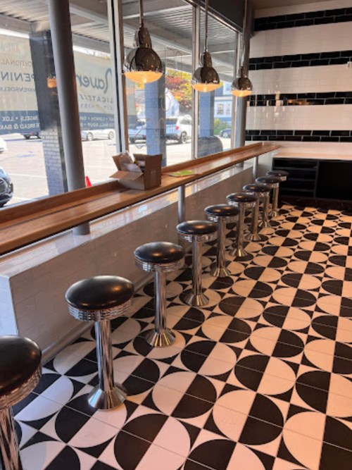 A modern café interior featuring a long wooden counter with chrome stools and a black and white patterned floor.