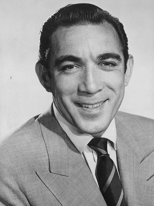 A smiling man in a suit and tie, with slicked-back hair, posed against a light background.