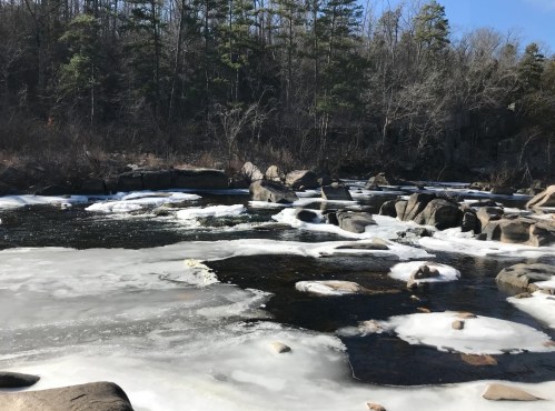 A serene river scene with ice and rocks, surrounded by trees in a winter landscape.