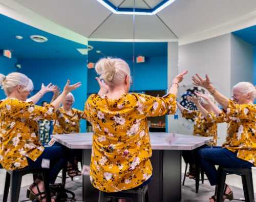 A woman with white hair in a floral yellow blouse gestures at a table, surrounded by mirrors reflecting her image.