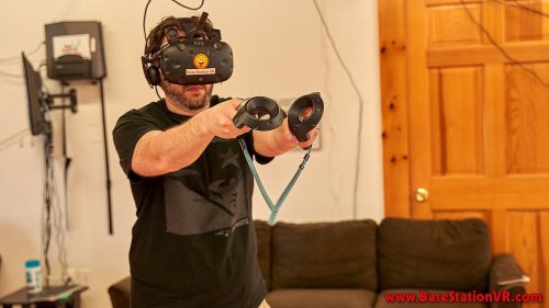 A person wearing a VR headset and holding controllers, standing in a room with a couch and wall-mounted equipment.