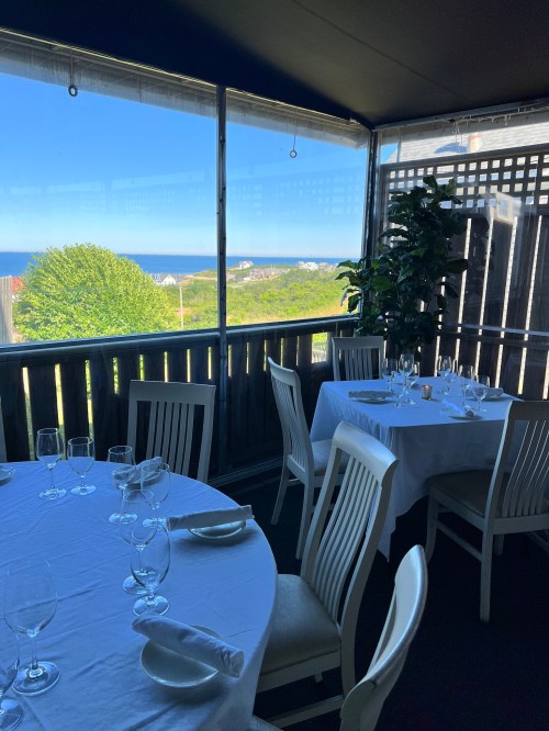 A dining area with white tablecloths and glasses, featuring a view of greenery and the ocean through large windows.