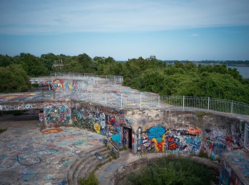 A graffiti-covered, abandoned structure surrounded by greenery and a view of the water in the background.