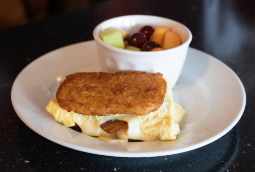 A plate with a cheesy omelet topped with a crispy hash brown and a side of mixed fruit in a bowl.