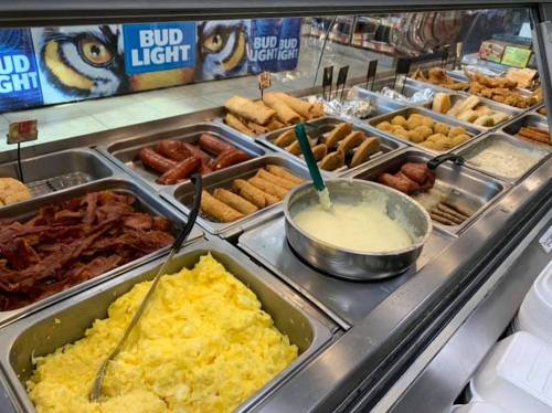 A buffet display featuring scrambled eggs, bacon, sausages, and various fried foods in metal trays.