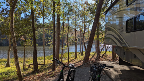 A serene lakeside scene with an RV parked among trees and two bicycles in the foreground.