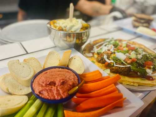 A plate with hummus, bread, celery, and carrots, alongside a bowl of salad and a pizza-like dish.