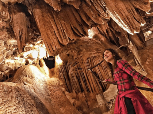 A woman in a red plaid shirt smiles while exploring a cave with impressive rock formations and warm lighting.