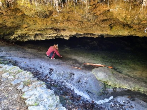 A person crouches by a rocky cave entrance, reaching into clear water with a stone path nearby.