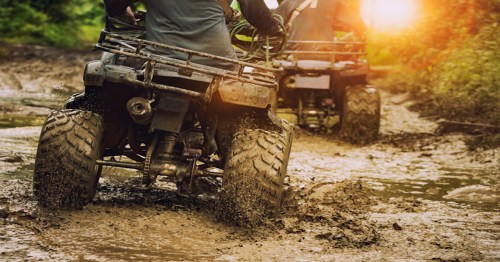 Two riders on ATVs navigate a muddy trail during sunset, with splashes of mud flying behind them.