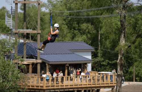 A person zip-lining above a wooden deck with onlookers, surrounded by trees and a building in the background.