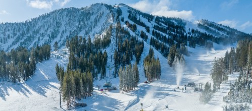 A snowy mountain landscape with ski slopes, trees, and snowmaking equipment under a clear blue sky.
