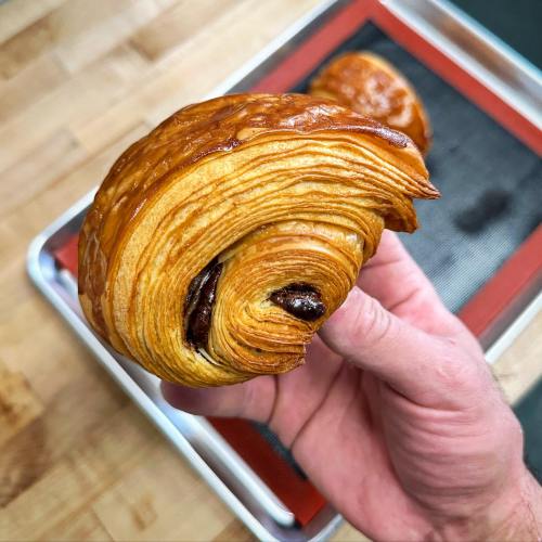 A hand holding a golden-brown, flaky pastry with a spiral shape and chocolate filling.