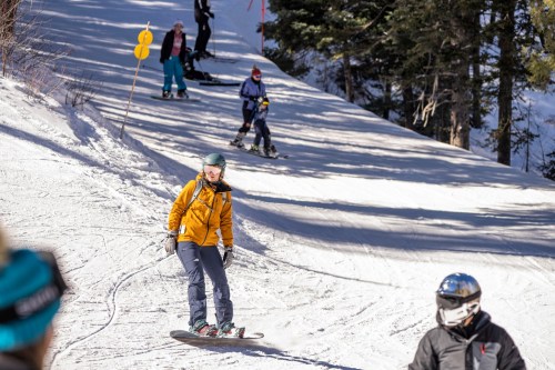 A skier in a yellow jacket glides down a snowy slope, with other skiers and snowboarders in the background.
