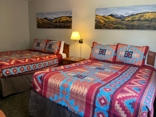 A cozy hotel room with two beds, featuring colorful patterned quilts and mountain landscape artwork on the walls.
