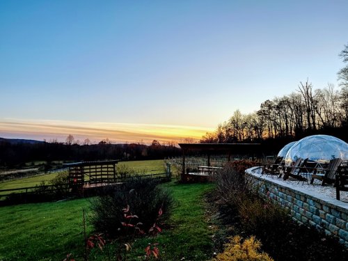 A serene landscape at sunset, featuring a dome structure, seating area, and lush green fields under a colorful sky.