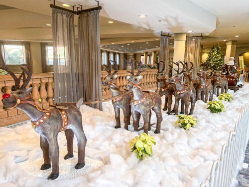 A row of decorative reindeer with red noses on a snowy display, with a Christmas tree and Santa in the background.
