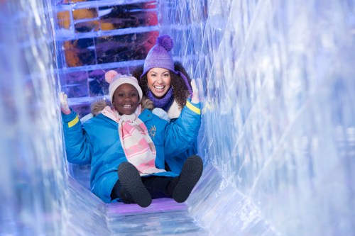 Two smiling women slide down an ice tunnel, wearing winter coats and hats, surrounded by colorful ice sculptures.
