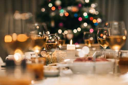 A festive dinner table with glasses of wine, candles, and a blurred Christmas tree in the background.