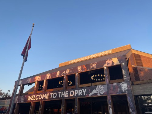 Exterior of the Grand Ole Opry House with a welcoming sign and a flag, under a clear blue sky.