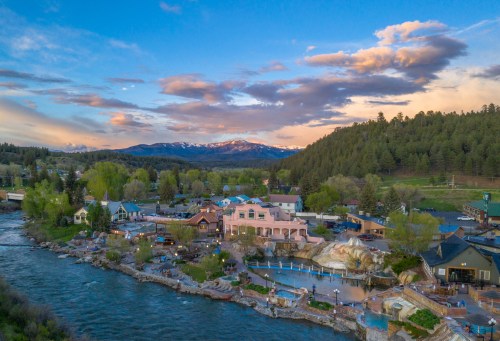 Aerial view of a scenic riverside town with mountains, lush greenery, and colorful buildings under a blue sky with clouds.