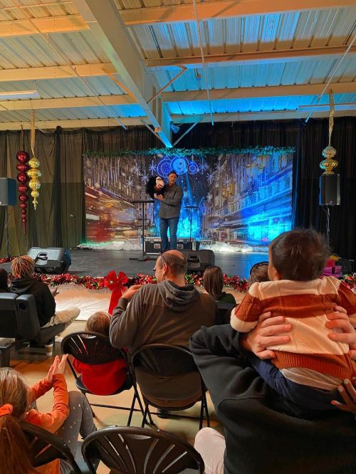 A performer on stage holds a child, while an audience watches from their seats, decorated for a festive event.