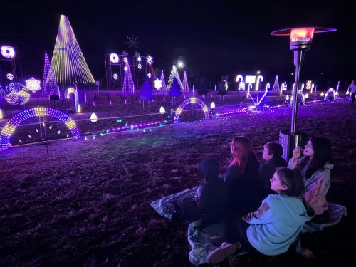 A group of people sits on a blanket, enjoying colorful holiday lights in a dark outdoor setting.