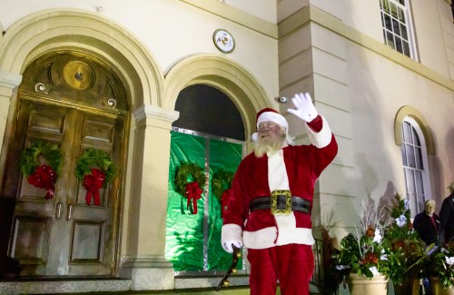 Santa Claus waves in front of a building decorated with wreaths and holiday lights.