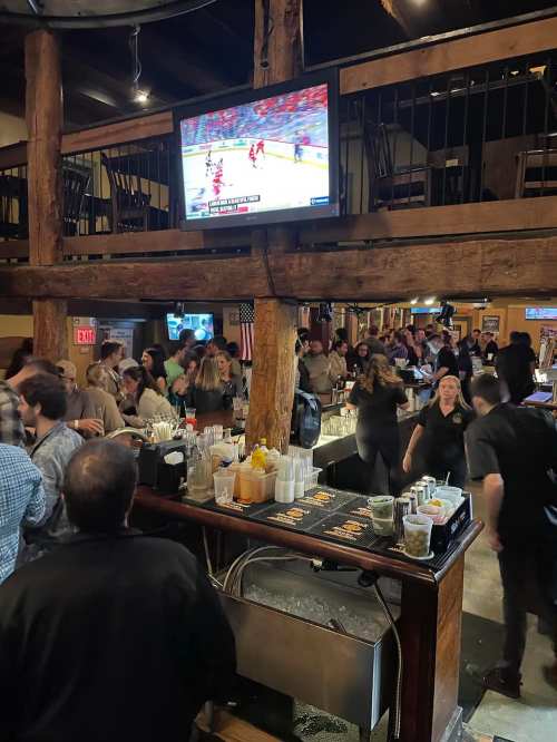 A busy bar scene with patrons socializing, a large TV showing a sports game, and a bartender serving drinks.