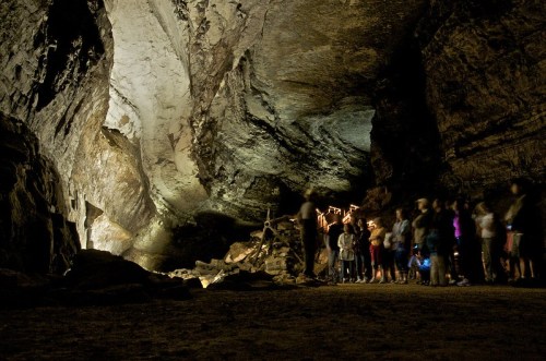 A group of people stands in a dimly lit cave, with rock formations and soft lighting creating an atmospheric scene.