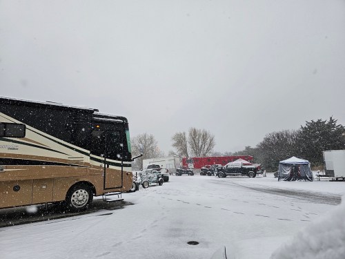 A snowy scene featuring an RV parked in a lot, with vehicles and tents in the background under falling snow.