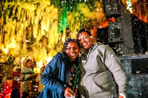 Two smiling women stand together in a festive setting with colorful lights and artificial snow falling around them.