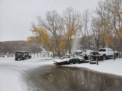 A snowy scene at a campsite with vehicles, trees, and a dusting of snow covering the ground and picnic tables.