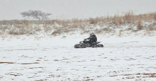 A person rides an ATV through a snowy landscape, with snowflakes falling and a sparse tree in the background.