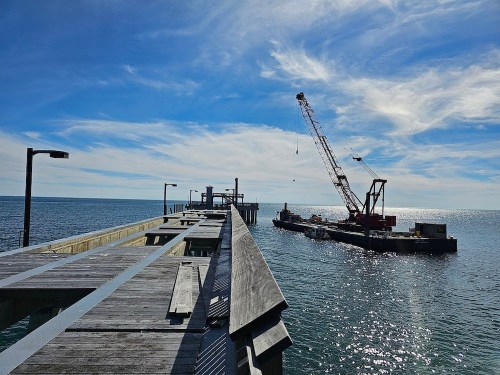 A wooden pier extends over calm water, with a crane barge working nearby under a blue sky with wispy clouds.