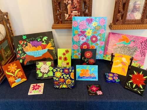 A colorful display of various floral and fruit-themed paintings arranged on a table against a wall.